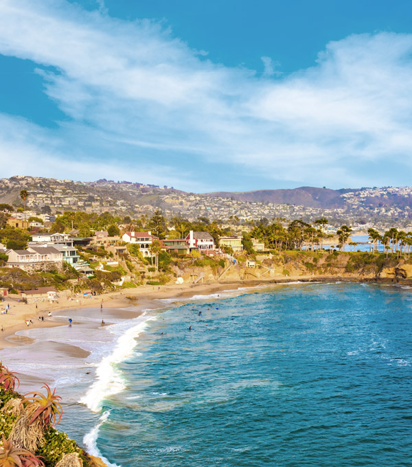 Rocky cliffs with palm trees fill the leftside foreground leading back to beach and breaking surf of Laguna Beach and city with hillside houses, California.