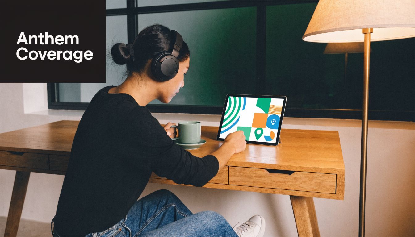 A person wearing headphones works on a tablet at a wooden desk with a lamp nearby.