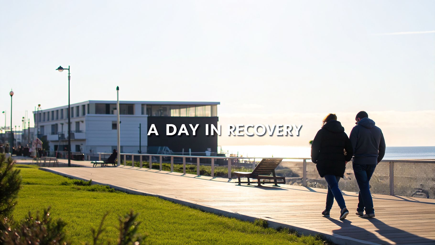 Two people walk along a sunny boardwalk near the ocean, with text 'A DAY IN RECOVERY' overlaid.