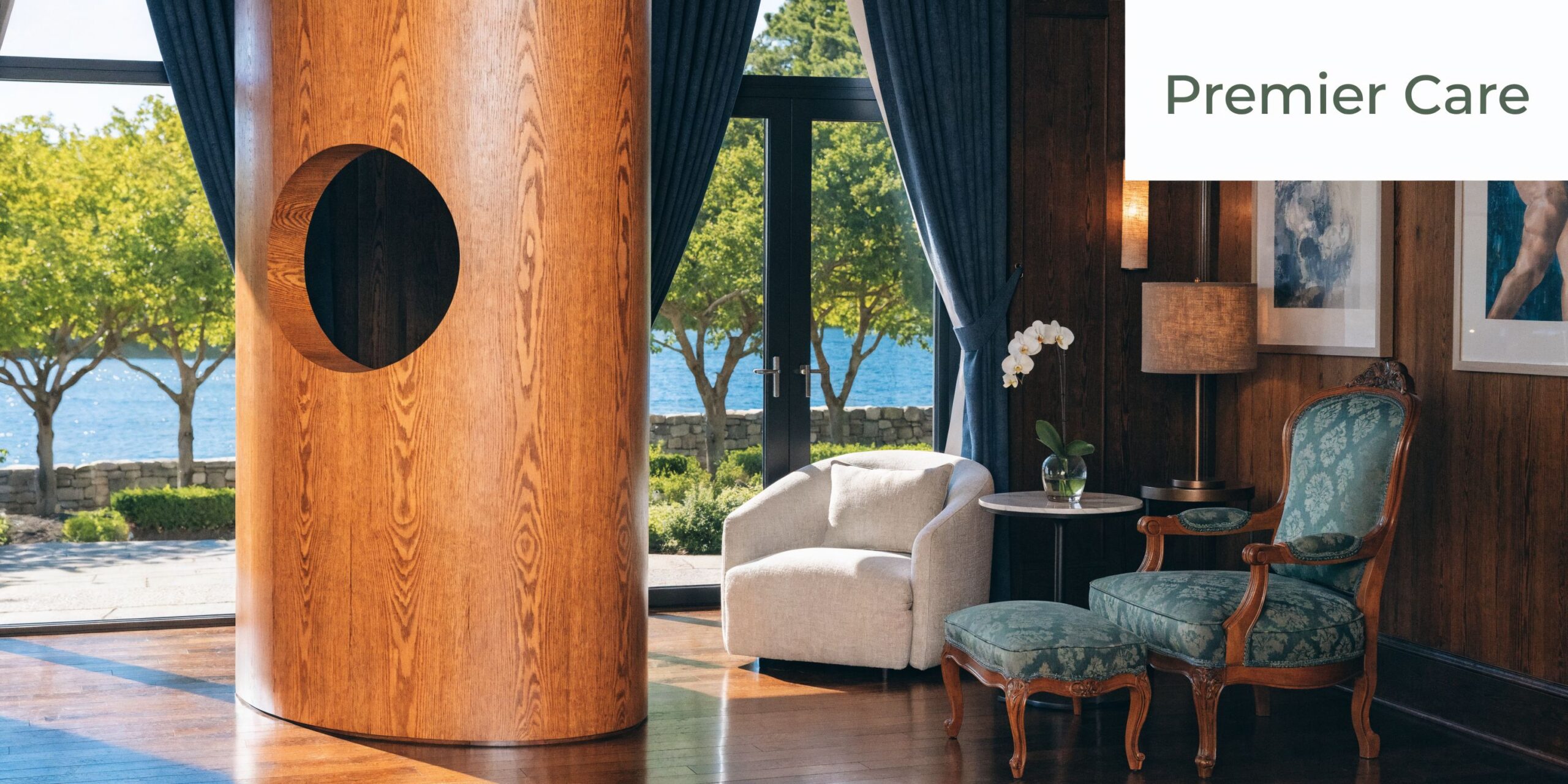 A luxurious sitting area with an elegant armchair, ottoman, and large wooden column overlooking a scenic waterfront.