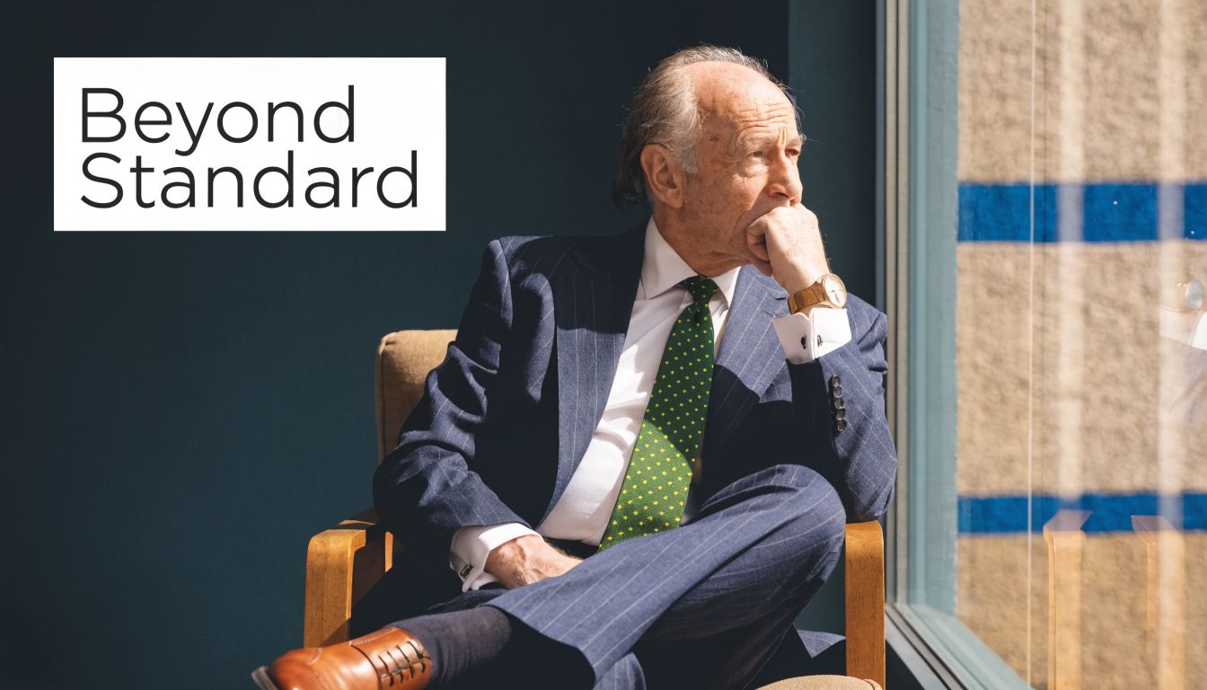 A sophisticated elderly businessman sitting in a chair, thoughtfully looking out a bright window.