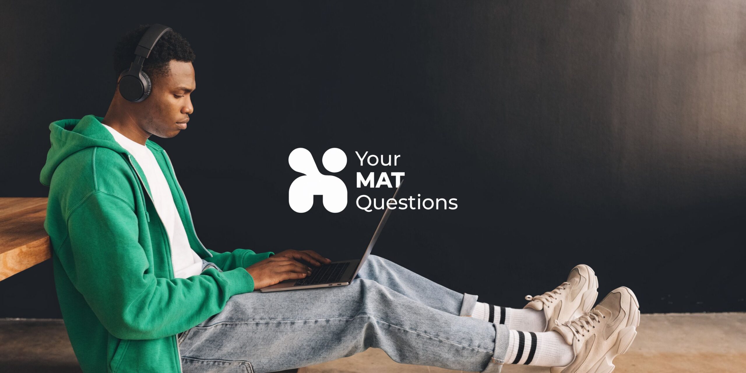 A young man wearing headphones and a green hoodie sits on the floor using a laptop computer.