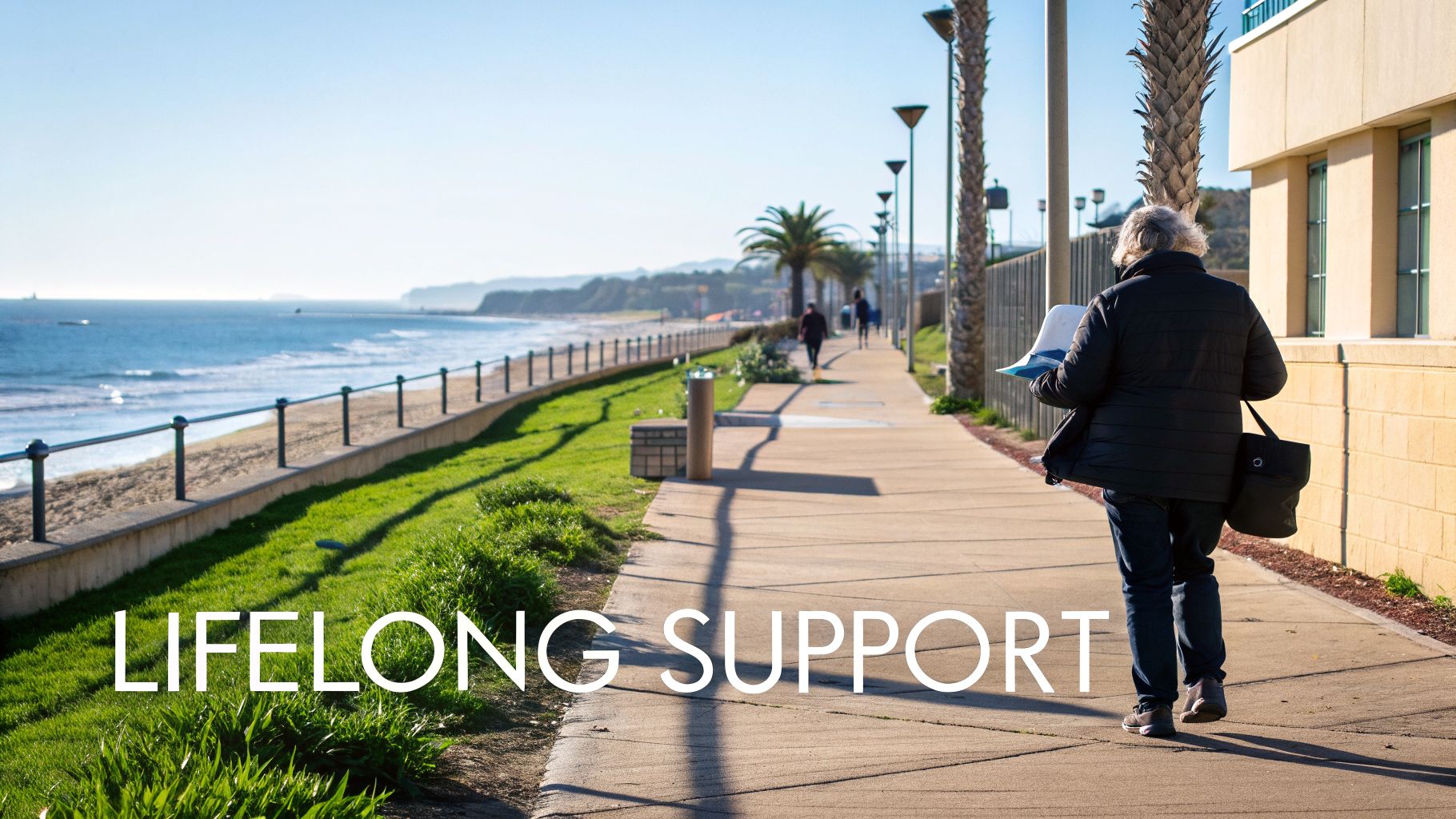 An older person walks along a sunny coastal promenade with the ocean, beach, and palm trees.