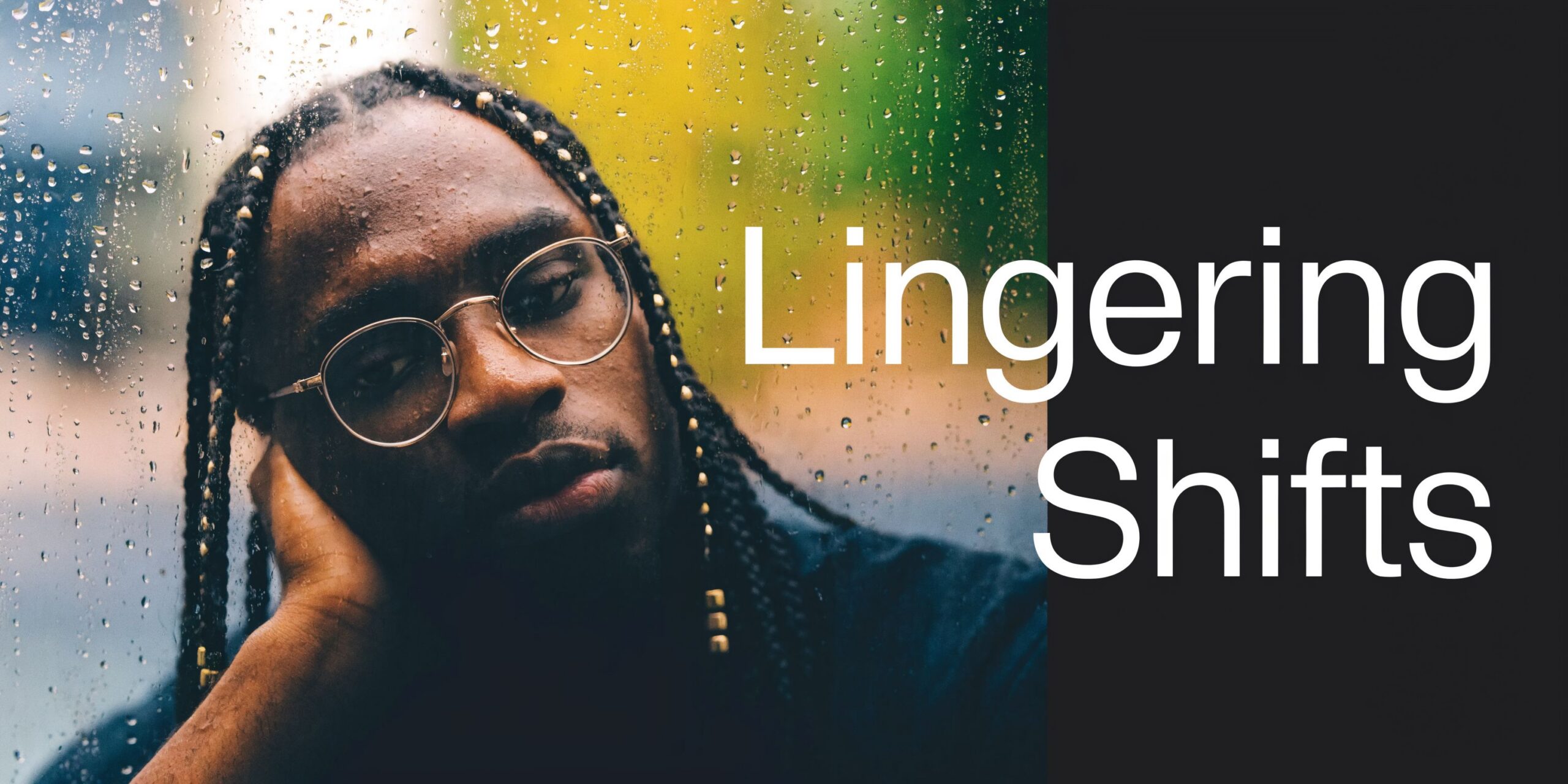 A pensive young man with braided hair and glasses looks through a window covered in raindrops.